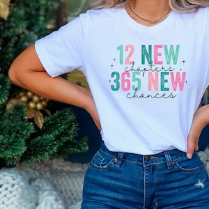 Op de afbeelding: Wit T-shirt met een groene en roze afbeelding met de tekst "12 Nieuwe Hoofdstukken + 365 Nieuwe Kansen".