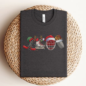 May include: A dark gray t-shirt with a Christmas-themed hockey design. The design features a hockey stick, skates, a hockey helmet, and a Santa hat.