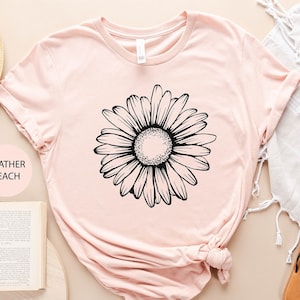 Puede incluir: Una camiseta de color melocot&oacute;n jaspeado con un gr&aacute;fico de margarita en blanco y negro. La camiseta est&aacute; colocada sobre una superficie marr&oacute;n claro con un sombrero de paja, un libro, un bolso tejido y unas sandalias.