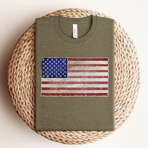 Pode incluir: Uma camiseta verde com um design de bandeira americana desbotada estampado na frente.