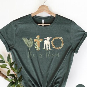 May include: A dark green t-shirt with a white lamb, a cross, a palm branch, and a crown of thorns. The text "He is Risen" is printed below the image.
