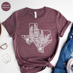 May include: Heather maroon t-shirt with a white graphic of the Texas state outline filled with floral designs. The shirt is styled with blue jeans and white sneakers. A wooden tray with a plant, camera, and accessories is also in the image.