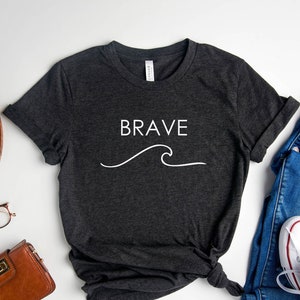 Op de afbeelding: Donkergrijs t-shirt met het woord "BRAVE" en een witte golfafbeelding. De afbeelding toont een blauwe jeans, witte sneakers, een bruine portemonnee en een vintage camera op een witte ondergrond.