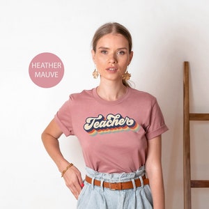 Puede incluir: Una camiseta de color malva jaspeado con el texto "Teacher's" en un dise&ntilde;o retro arco&iacute;ris.