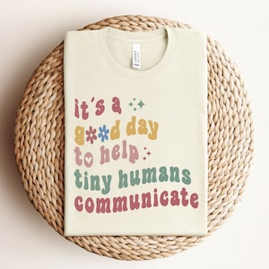 Puede incluir: Una camiseta beige claro con el texto "it's a good day to help tiny humans communicate" en letras retro de colores.