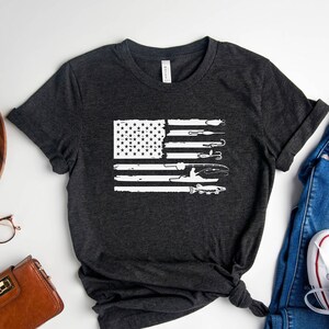 K&ouml;nnte beinhalten: Ein dunkelgraues T-Shirt mit einem wei&szlig;en, abgenutzten Design einer amerikanischen Flagge mit Werkzeugen. Die Flagge besteht aus Werkzeugen wie Schraubenschl&uuml;sseln, H&auml;mmern und Schraubendrehern.