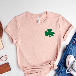 May include: A pink t-shirt with a green shamrock design. The shirt is laid out on a white surface with a pair of jeans, a book, a pair of glasses, and a small brown wallet.