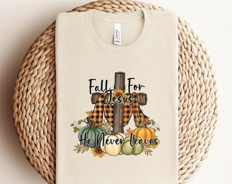 May include: A beige t-shirt with a fall-themed design featuring a wooden cross with a plaid scarf, sunflowers, and pumpkins. The text "Fall For Jesus He Never Leaves" is printed on the shirt.