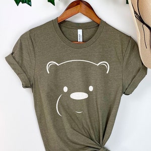 May include: A green t-shirt with a white bear face graphic. The shirt is knotted at the bottom and hanging on a wooden hanger.