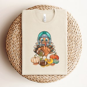 May include: A white t-shirt featuring a cartoon image of a turkey wearing a teal football helmet and holding a football. The turkey is surrounded by pumpkins and autumn leaves.