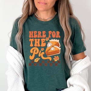 May include: A green t-shirt with the text "Here for the Pie" in a retro font. The shirt features a graphic of a slice of pumpkin pie with whipped cream and fall leaves.