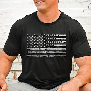 May include: Black t-shirt with a distressed American flag design. The flag has the words "Husband, Daddy, Protector, Hero" written on it.