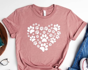 May include: A mauve t-shirt with a white heart design made of paw prints. The shirt is laid out on a white surface with a pair of blue jeans, white sneakers, a wooden tray with a plant and a camera, and a brown wallet and glasses.