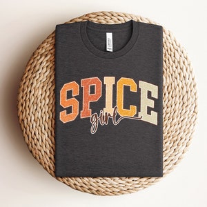 May include: A dark gray t-shirt with the text "SPICE girl" in a retro font with orange, yellow, and brown colors.