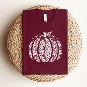 May include: A maroon t-shirt with a white, decorative pumpkin design. The pumpkin is filled with a swirling, floral pattern.