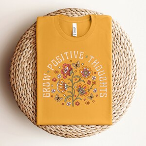 May include: A mustard yellow t-shirt with a floral design and the text "Grow Positive Thoughts" in a circle around the flowers.