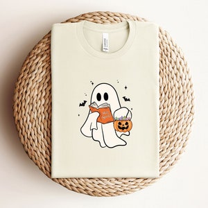 May include: A cream-colored t-shirt with a cartoon ghost holding a book that says "Scare Spook" and a pumpkin bucket filled with candy.