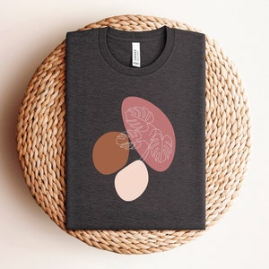 Puede incluir: Una camiseta gris oscuro con un dise&ntilde;o gr&aacute;fico de formas abstractas en tonos de rosa, marr&oacute;n y blanco. El dise&ntilde;o presenta una hoja estilizada con un dibujo lineal de una planta.