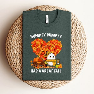 May include: A dark green t-shirt with a white graphic of a cartoon egg with a scarf and a cup of coffee. The egg is standing next to a wagon with pumpkins. The text "Humpty Dumpty Had A Great Fall" is printed on the shirt.