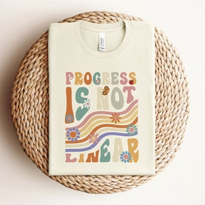 May include: A beige t-shirt with a retro-style graphic that says "Progress is not linear" in colorful, wavy letters. The graphic features flowers and butterflies.