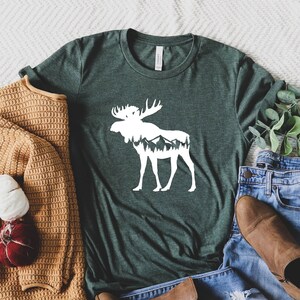 May include: A dark green t-shirt with a white moose graphic, mountains inside the moose silhouette, and a crew neck. The shirt is paired with blue jeans, brown boots, and a tan sweater. A decorative bundle of yarn and a red ornament are also included.