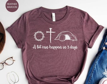 Puede incluir: Camiseta color borgoña con un gráfico blanco de una corona de espinas, una cruz y una tumba vacía. El texto "A lot can happen in 3 days" está debajo. La camiseta se combina con vaqueros azules y zapatillas blancas.