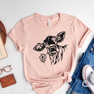 May include: A pink t-shirt with a black silhouette of a cow with a flower in its mouth. The shirt is laid out on a white surface with a pair of jeans, a book, a plant, glasses, and a wallet.