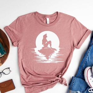 K&ouml;nnte beinhalten: Ein mauvefarbenes T-Shirt mit einer wei&szlig;en Silhouette von Arielle, der kleinen Meerjungfrau, die auf einem Felsen vor einem Vollmond sitzt, mit einer Reflexion im Wasser.