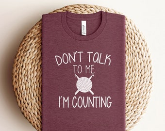 May include: A maroon t-shirt with the text "Don't talk to me I'm counting" and a white illustration of yarn and knitting needles.