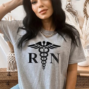 May include: Heather grey t-shirt with a black medical symbol and the letters "RN" printed on the front.
