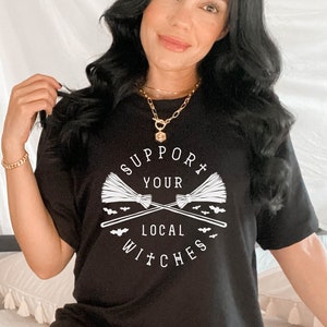May include: Black t-shirt with a white graphic that says "Support Your Local Witches" with crossed broomsticks and bats.