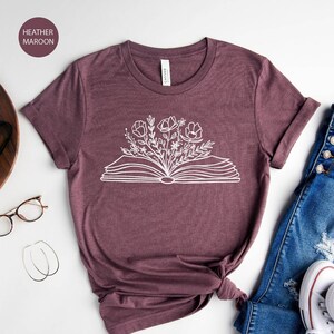 K&ouml;nnte beinhalten: Ein T-Shirt in Heide-Maroon mit einer wei&szlig;en Grafik eines offenen Buches, aus dem Blumen wachsen. Das Shirt liegt auf einer wei&szlig;en Oberfl&auml;che mit einer Brille, einer Kamera, einer Pflanze, einer Jeans und wei&szlig;en Turnschuhen.
