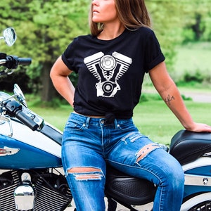 May include: A woman wearing a black t-shirt with a white motorcycle engine graphic sits on a blue and white motorcycle. She is wearing blue jeans with rips and a tattoo on her arm.