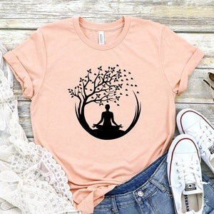 May include: A peach-colored t-shirt with a black silhouette of a person meditating under a tree with leaves. The tree is inside a circle. The t-shirt is on a wooden surface with a white lace shawl, denim shorts, and white sneakers.