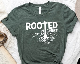 Può includere: Una maglietta verde con il testo "Rooted in Christ" e una grafica bianca di un albero con radici e una croce.