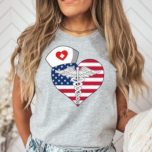Puede incluir: Una camiseta gris con un diseño de corazón rojo, blanco y azul. El corazón presenta un símbolo de caduceo médico y un gorro de enfermera con una cruz roja.
