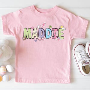 May include: A pink t-shirt with the name "MADDIE" in colorful, Easter-themed letters. The shirt is next to white sneakers, a plush bunny, and decorative Easter eggs.