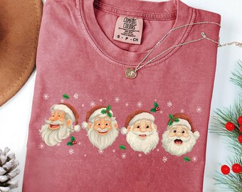 May include: A dusty rose Comfort Colors t-shirt featuring four Santa Claus faces with white beards and red hats. The shirt has a crew neck and a small tag. A silver necklace with a pendant is draped across the shirt.