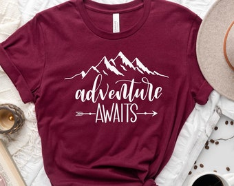 May include: A maroon t-shirt with white text that reads "adventure awaits" with a mountain range graphic above the text and arrows pointing to the left and right of the text.