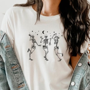 May include: White t-shirt featuring a graphic of three skeletons dancing under a crescent moon and stars.