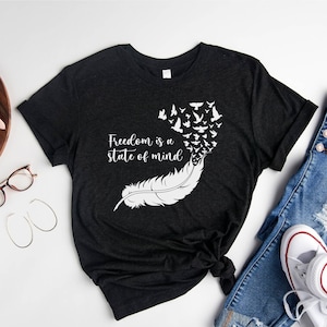 May include: A dark gray t-shirt with a white feather design and the text "Freedom is a state of mind". The feather has a flock of birds flying out of it.