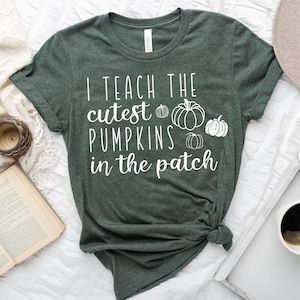 Pode incluir: Uma camiseta verde com o texto "Eu ensino as abóboras mais fofinhas do pomar" em letras brancas. Há quatro pequenas ilustrações de abóboras brancas na camiseta.