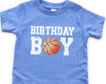 Può includere: Una maglietta blu a maniche corte con il testo "BIRTHDAY BOY" in bianco e una grafica di pallacanestro al centro.