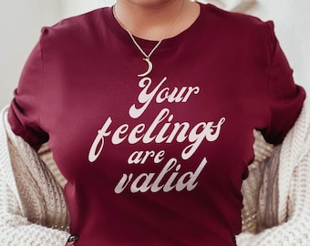 Op de afbeelding: Een maroon t-shirt met de tekst "Your feelings are valid" in een roze retro-lettertype.