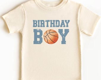 Può includere: Una maglietta color crema con una grafica di pallacanestro blu e arancione e il testo "Birthday Boy".