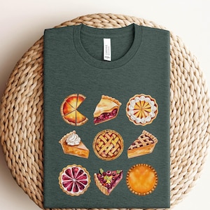 May include: A dark green t-shirt featuring a design of nine different pie slices. The pies are all various flavours, including apple, cherry, and pumpkin. The design is in a watercolour style.