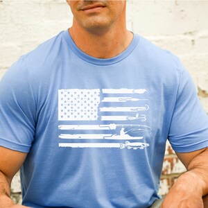 K&ouml;nnte beinhalten: Ein hellblaues T-Shirt mit einem wei&szlig;en, abgenutzten Design der amerikanischen Flagge. Die Flagge zeigt fischereibezogene Bilder, darunter einen Fischer in einem Boot, Angelruten, Haken und Fische.