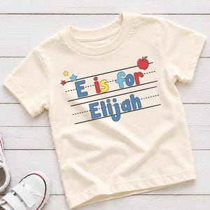 May include: A cream-colored short-sleeve t-shirt featuring the text E is for Elijah printed in blue, yellow, and red letters on black lined paper graphics. A small red apple and tiny blue and yellow stars are positioned above the text.