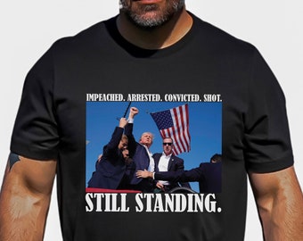 May include: Black t-shirt with a white graphic of a man holding a flag and the text "Impeached. Arrested. Convicted. Shot. Still Standing."