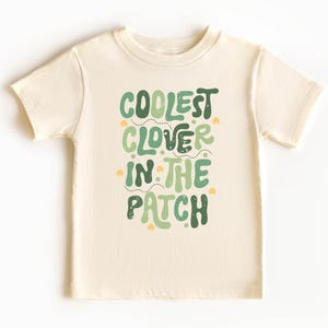 Könnte beinhalten: Cremefarbenes T-Shirt mit dem Text "COOLEST CLOVER IN THE PATCH" in Grüntönen. Das Design beinhaltet Kleeblatt-Grafiken. Das Shirt hat einen Rundhalsausschnitt und kurze Ärmel.
